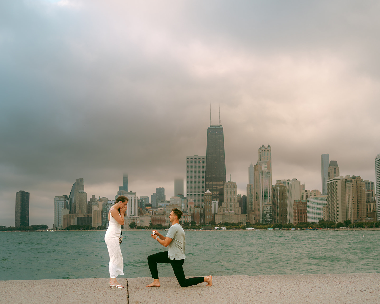Man kneeling to propose at North Avenue Beach against a stormy Chicago skyline with the Hancock in cloud.